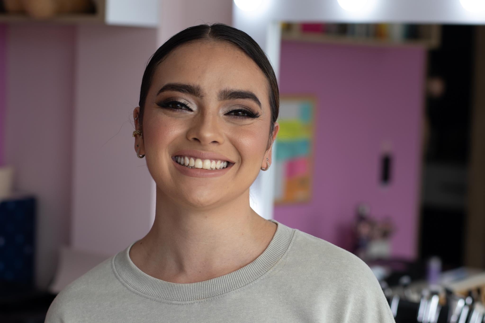 Retrato sonriente con maquillaje de ojos ahumado y cabello recogido, en un estudio de paredes rosas