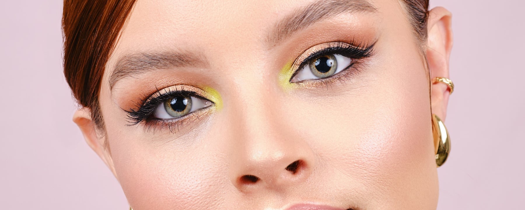 Primer plano de los ojos con sombra dorada y verde, delineado alado y pestañas, piel luminosa sobre fondo rosa