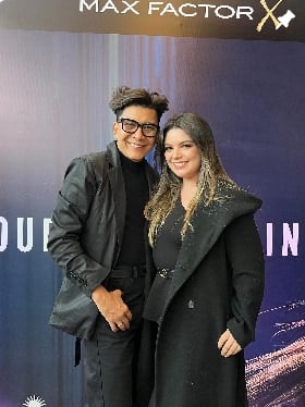 Dos personas posan frente al panel de un evento de Max Factor: él con chaqueta de cuero negra y ella con abrigo largo negro y cabello rubio ondulado