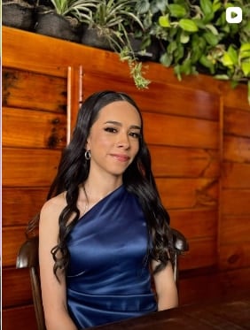 Mujer de cabello largo ondulado con maquillaje suave y un vestido azul de satén de un solo hombro, sentada junto a una pared de madera con plantas
