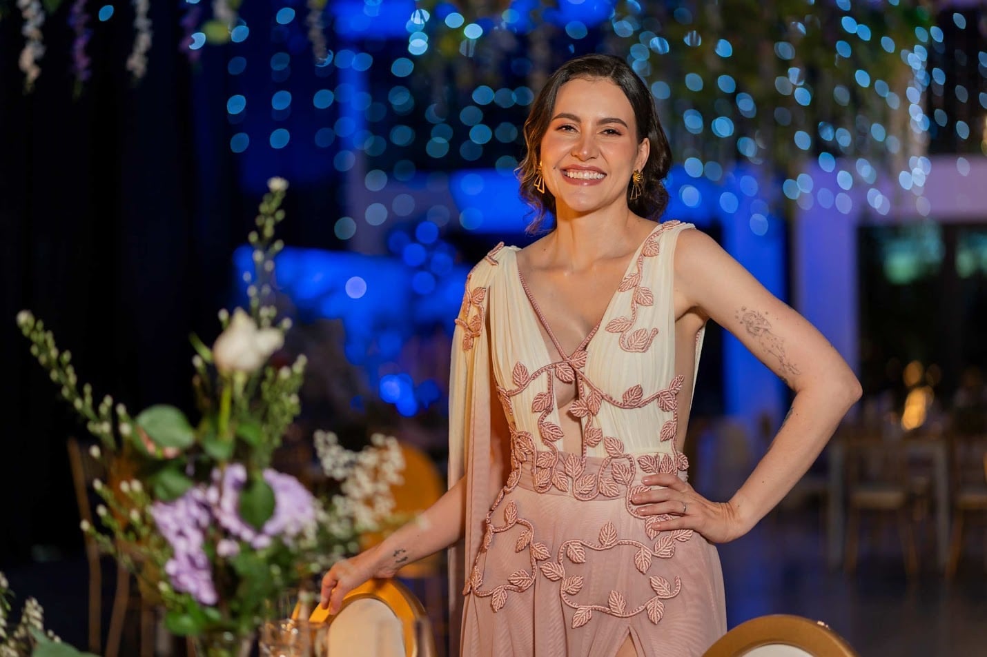 Maquillaje social para eventos de noche: mujer sonriente con vestido rosa y crema con apliques de hojas y las manos en la cintura, con luces azules de fondo