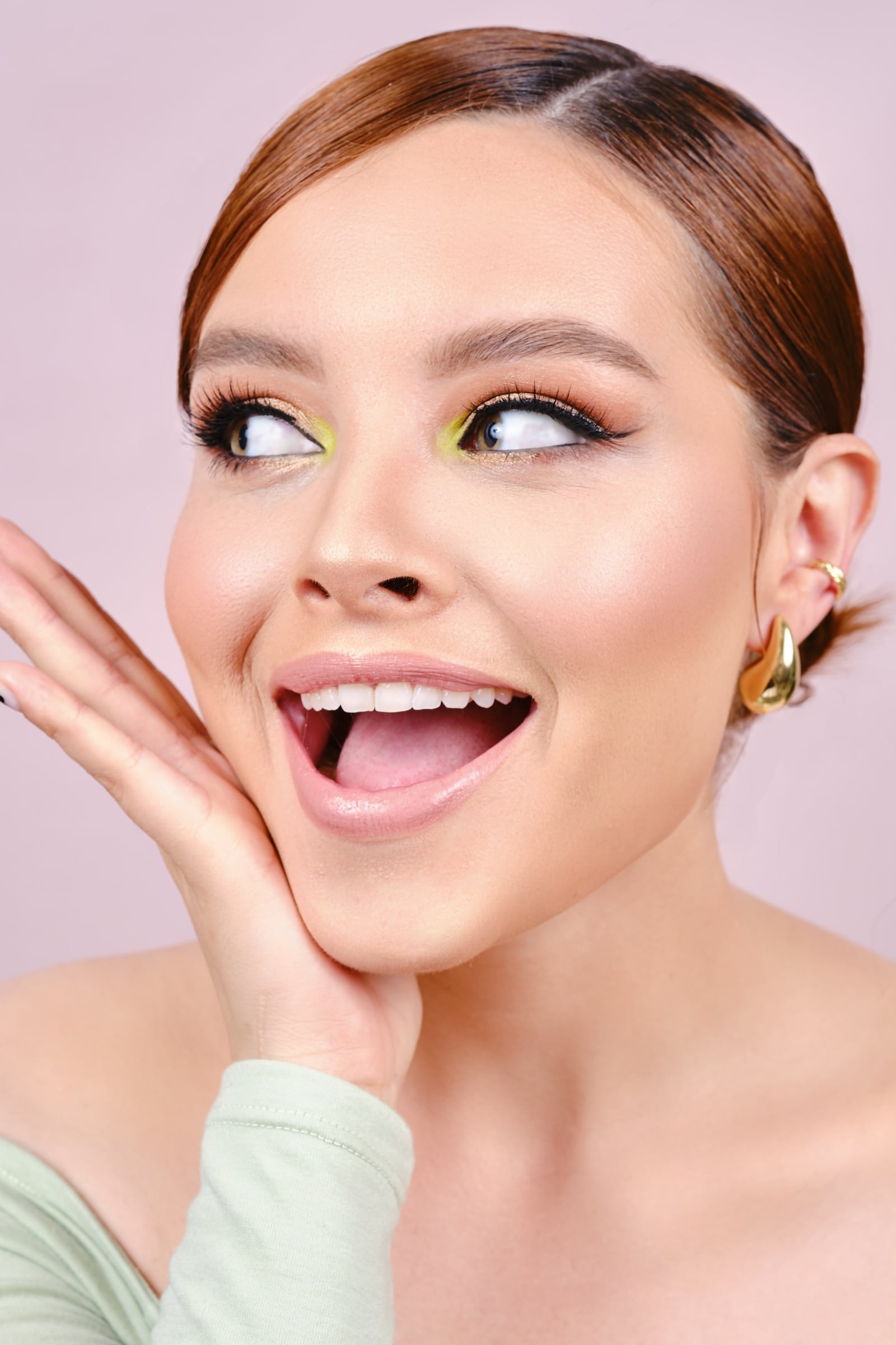Maquillaje editorial con sombra verde y amarilla y delineado alado: mujer de cabello recogido con una mano junto a la mejilla, sobre fondo rosa
