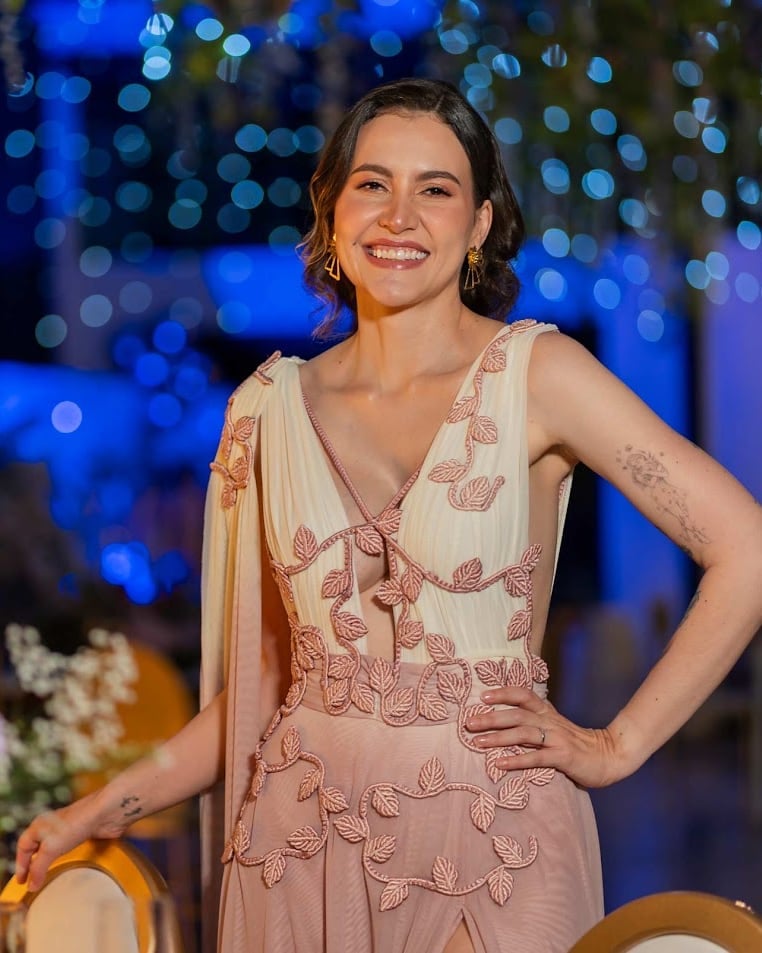 Mujer sonriente con vestido rosa y crema con apliques de hojas y las manos en la cintura, en un evento de noche con luces azules